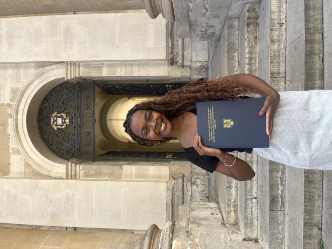 a young woman holding a bound copy of her dissertation on the steps of a grand stone building