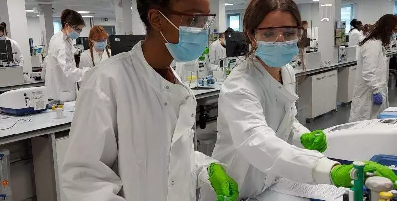two students working at a lab bench with lab coats, gloves and masks